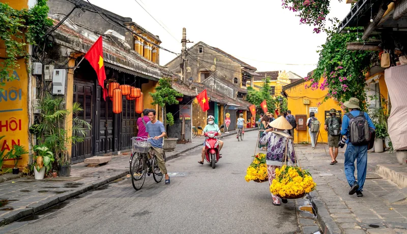 A street vendor carries baskets of yellow flowers past yellow colonial buildings in Hoi An's old town.