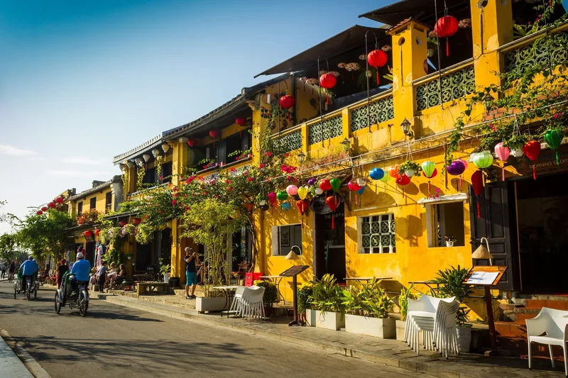 Colorful lanterns decorate yellow facades along a quiet street in Hoi An's old town as cyclists pass by.