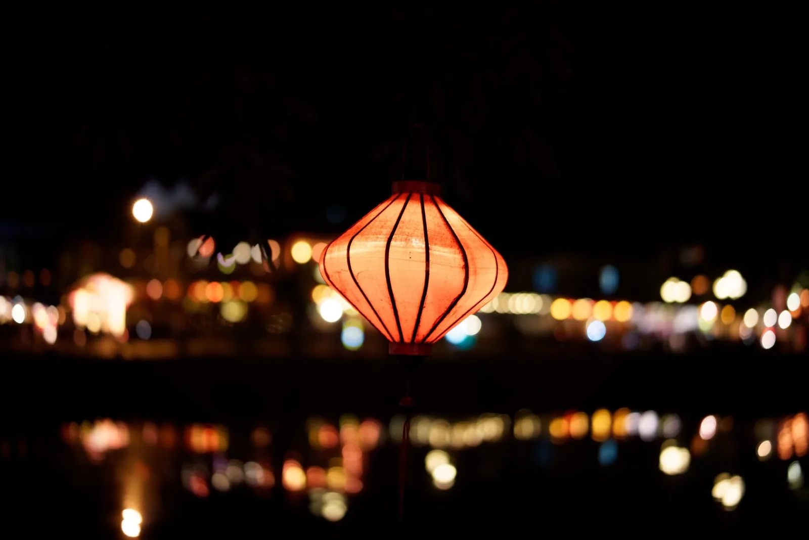 A glowing red silk lantern hangs over the river at night with bokeh city lights in Hoi An.