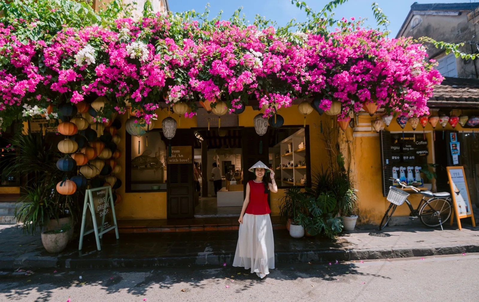 A woman in a conical hat stands before a bougainvillea-covered yellow shophouse in Hoi An.