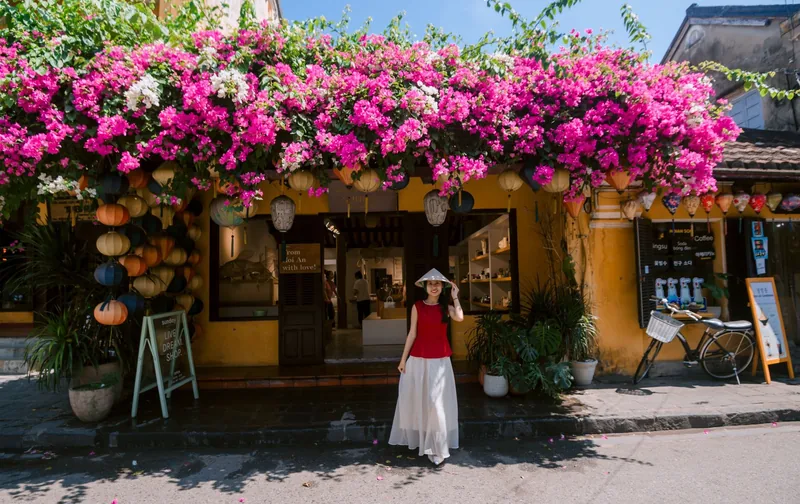 A woman in a conical hat stands before a bougainvillea-covered yellow shophouse in Hoi An.