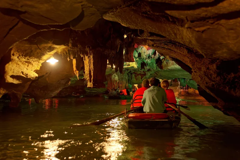 Tourists in life jackets row a small boat through an illuminated limestone cave at Trang An, Ninh Binh.