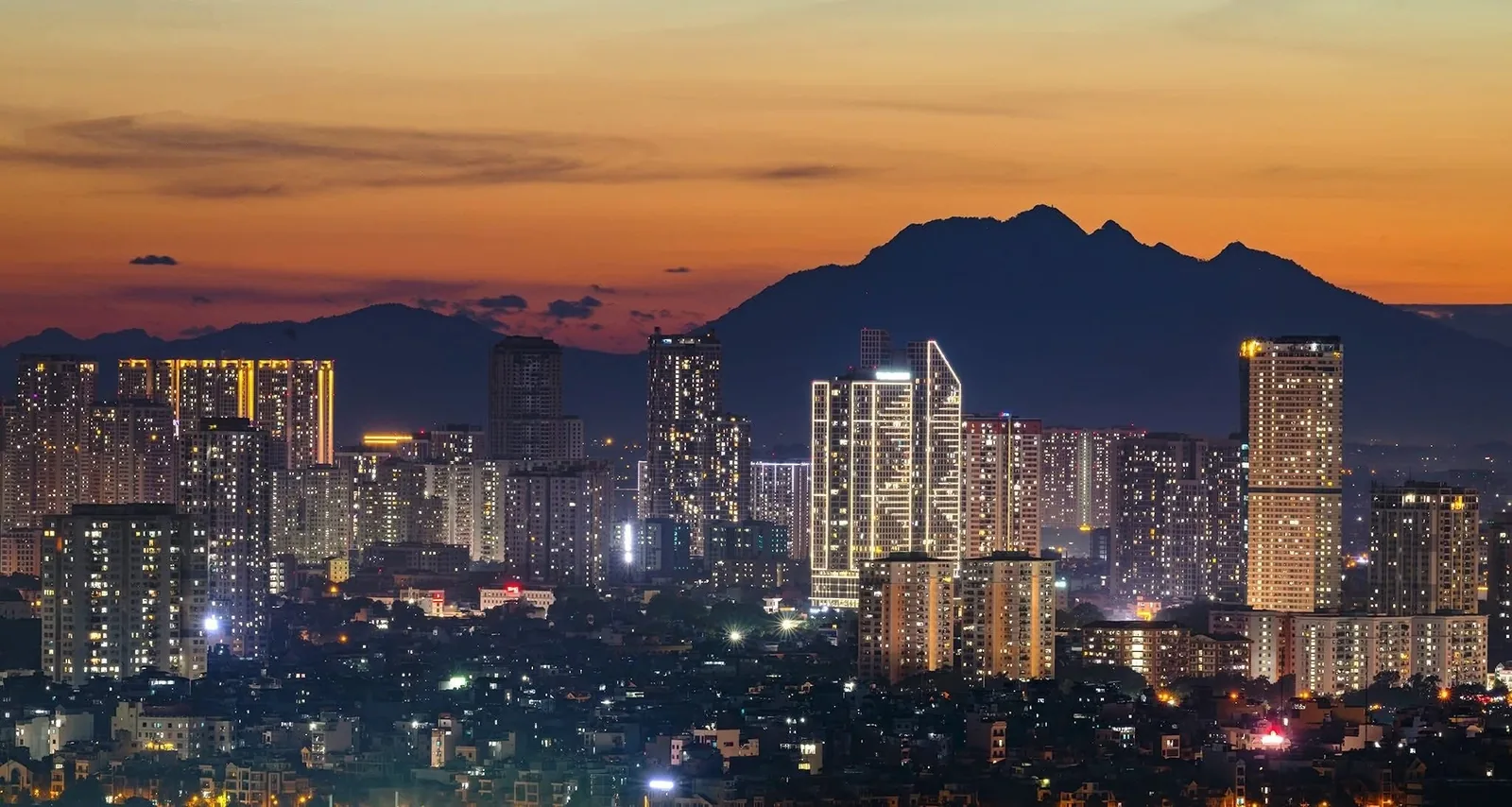City skyline of Hanoi glowing at dusk with Ba Vi mountain silhouetted against an orange sky
