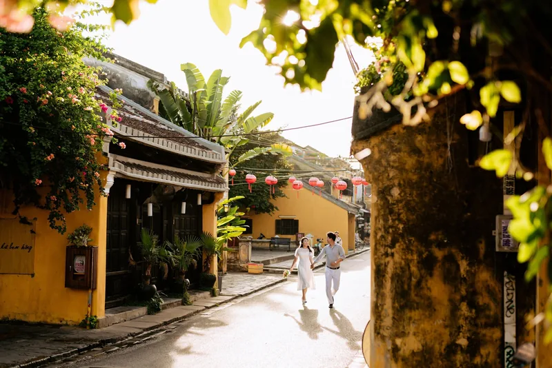 A couple holds hands walking down a sunlit lantern-lined street in Hoi An's old town.