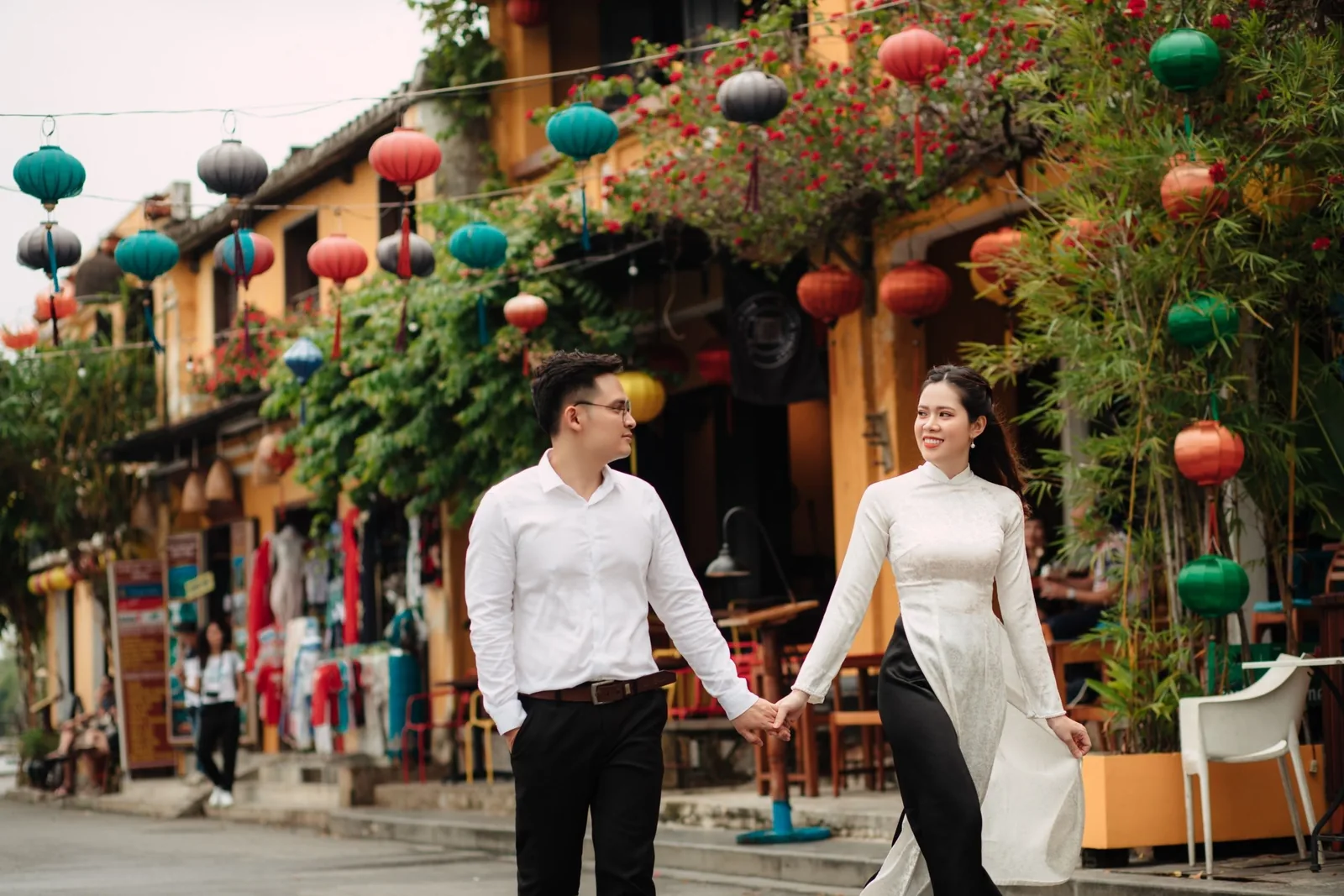 A couple hold hands walking past colorful lanterns and yellow buildings on a street in Hoi An.